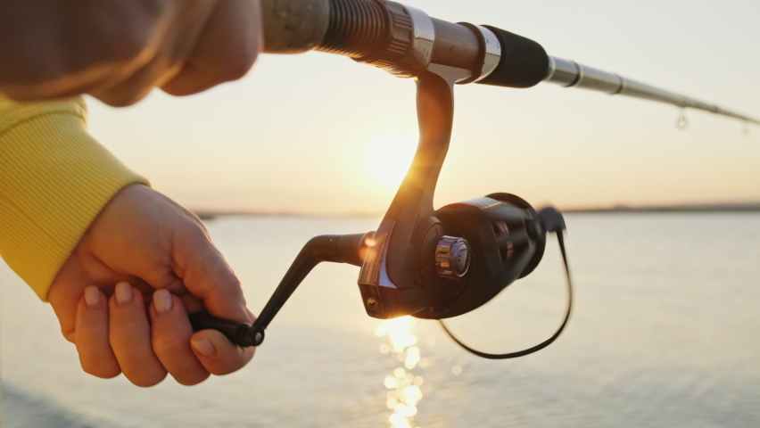 Reel of fishing rod hand rotation of orange sunset slow motion. Woman hobby fishing on sea tightens fishing line reel fish summer. Lens flare. Calm surface sea. Bright disk of sun Go Everywhere Royalty-Free Stock Footage #1059863699