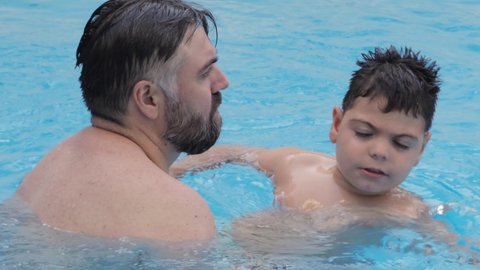 Caucasian child with disability, reduced mobility, swims, relaxes and plays with his father, a bearded man, in a pool during the summer.