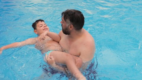 Caucasian child with disability, reduced mobility, swims, relaxes and plays with his father, a bearded man, in a pool during the summer.