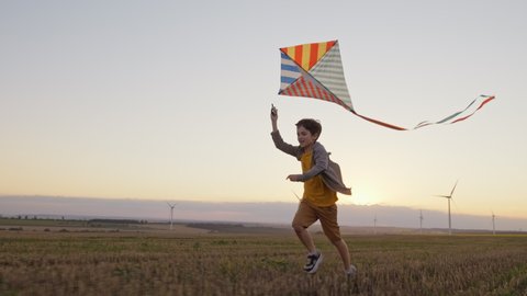 Happy boy runs wheat field launches bright kite into sky mown playing with wind in field of an orange sunset on day lens flares wind turbines in summer slow motion. School break. Lifestyle. Childhood