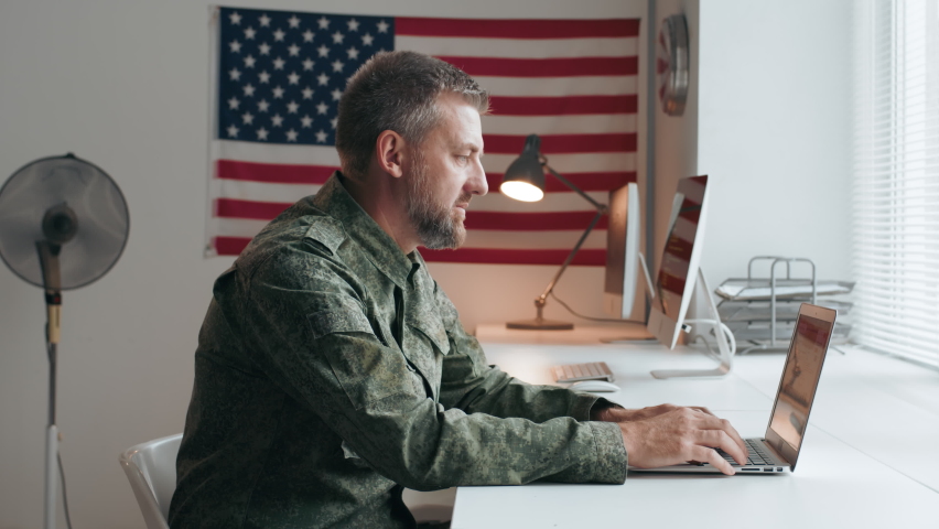 Portrait of middle-aged male U.S. army officer in military uniform sitting at desk and typing on laptop, then turning towards camera and posing