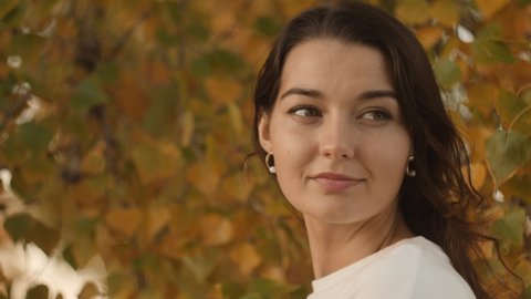 Portrait of a romantic brunette woman standing in the park at fall on a sunny day smiling and looking away. The face of a happy beautiful woman in autumn outdoor at sunset. Slow-motion 4k footage