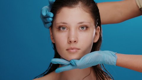 doctor in blue gloves moving woman head isolated on blue, pretty brunette with clean skin checking head before operation by beautician