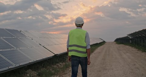 Backside view of male engineer in uniform walking and looking at solar power plant. Man in hard helmet examining object. Concept of solar station development and green energy