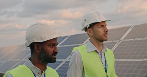 Close up of diverse male engineers walking at solar farm and discussing efficient plan of construction. Two men in protective helmet and uniform talking. Concept of alternative energy