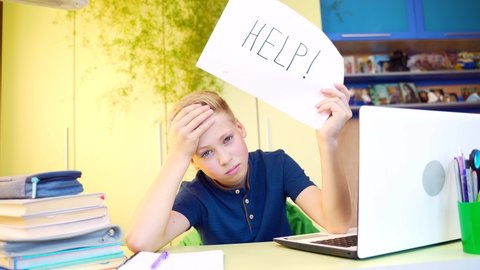portrait of a tired young blond boy teaches lessons with a notebook and laptop at table in the home. Teen schoolboy shows a picture with the inscription help while studying. Close-up face exhausted