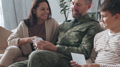 Slowmo medium shot of happy young woman and boy sitting on couch with male army veteran in uniform and listening to him share stories while looking at photos
