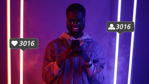 Portrait of happy African American guy blogger standing in neon lights and texting on smartphone. Handsome joyful male influencer with user interface. Social media likes and followers growth icons