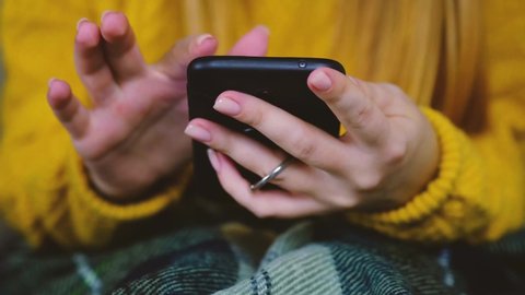 Young female hands touch the phone. Woman in a yellow sweater covered herself with a green plaid blanket with a smartphone. Tapping, scrolling, watching video, content, bloggs. Modern lifestyle