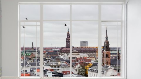 Copenhagen cityscape as seen from a white window while birds are flying. It is seen the city Christiansborg Palace and City Hall Square during a cloudy day. Copenhagen province, Denmark 