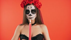 Day of Dead. Creepy lady with skull make-up, in black dress and headband of roses. She rasing up burning candle, sharply extinguishing it and smiling. Red background. Halloween. Close up, copy space - Powered by Shutterstock - Get 15% off with code: PIKWIZARD15