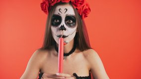 Day of Dead. Angry female with skull make-up, in black dress and headband of roses. Holding burning candle, looking at it and extinguishing, posing on red background. Halloween. Close up, copy space - Powered by Shutterstock - Get 15% off with code: PIKWIZARD15