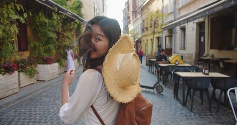 Rear of happy pretty Asian female with hat walking in old town outdoors in good mood. Close up of cheerful young woman traveler smiling on street in city. Urban tourism. Travel concept: stockvideo