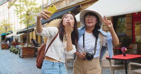 Cheerful Asian mother and daughter making selfie photo on smartphone on street. Portrait of happy female tourists posing and waving hands while taking pictures on cellphone in city. Travel concept