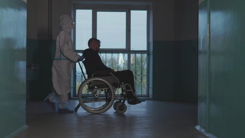 A doctor in a medcial protective coverall pushes a wheelchair with a patient in front of him in a dark corridor of a hospital