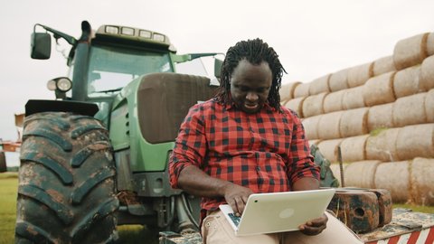 Thumb up. Young african farmer using tablet while sitting on green tractor. Haystack in the background. High quality 4k footage