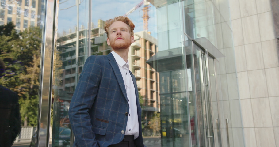 Young guy with red beard in checkered jacket and white shirt stands by mirror building wall reflecting city street at sunlight