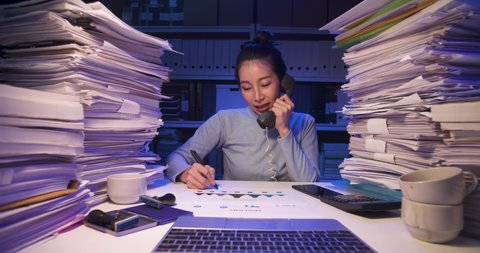Asian business woman sitting at desk covered with stack of paperwork and talking phone, using laptop. Tired alone girl sleeping while working hard at late night. Time lapse, Fast speed, Web cam view
