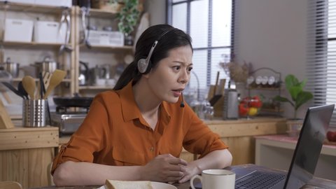furious woman manager wearing headset having a con-call or phone conversation from computer is talking loudly with hand gestures in the dining room.