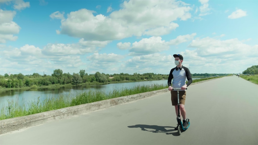 A man in a cap during sports on a scooter on the road near the river takes off a medical mask and greets someone. High quality footage. N1H1 coronavirus quarantine.