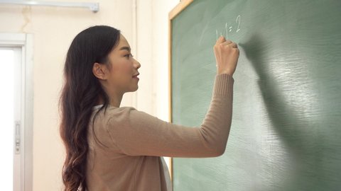 Young woman using chalk on chalkboard, education in school classroom. Female Asian teacher writing mathematics sums on blackboard