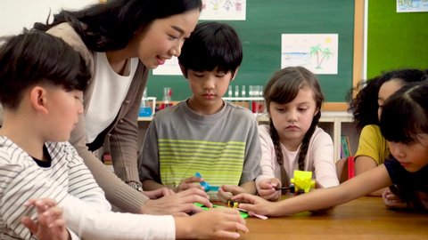 Young woman working in school helping multiethnic children make toy cars, learning about mechanics, stem. Asian school teacher assisting elementary students in science classroom