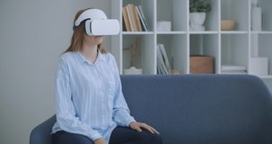 Young cheerful woman wearing virtual reality headset watching 360 VR video movie sitting in the bed at home. Portrait Of Young Woman In Virtual Reality Glasses Sitting On The Sofa At Home.