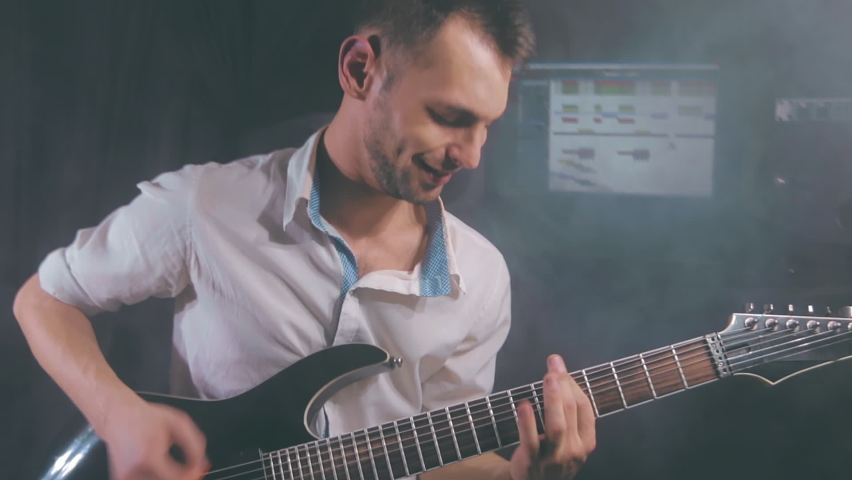 Medium Close-Up Shot of a Handsome Young Man Playing Electric Guitar in a Dark Smoky Room. Active Emotional Musician in a White Shirt is Recordind Music at the Home Studio Against the Black Background