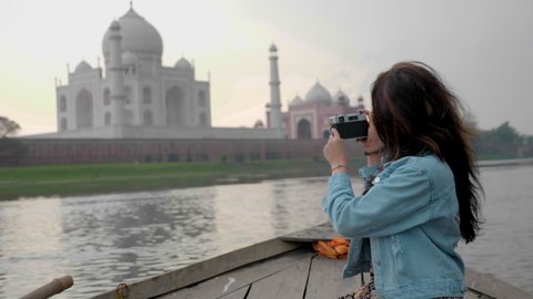 A young beautiful girl or female is sitting on a wooden rowing boat taking a picture of the Taj Mahal Mausoleum next to the Yamuna river. A woman solo traveller on holidays is clicking a photo on DSLR