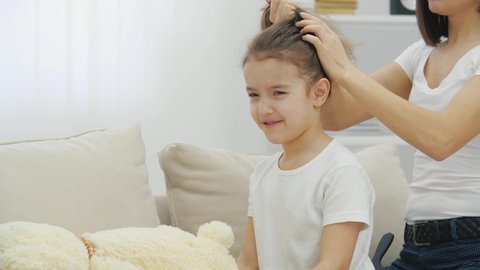 Girl's hair is tousled. Mother is trying to tie a ponytale, but as she pulling hair, daughter is almost crying.