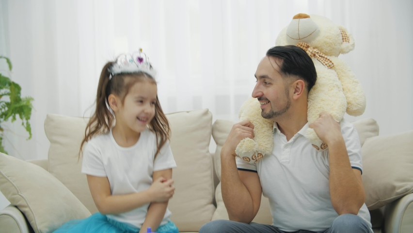 Cute princess girl making her father beautiful, wearing him in crown and necklace. Father is holding teddy bear on shoulders.