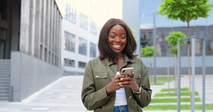 African American joyful young stylish woman tapping or scrolling on smartphone and standing at city street. Beautiful happy female texting message on mobile phone and smiling. Outside.