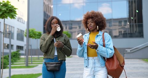 African American young pretty females, best friends talking cheerfully and walking with cups of coffee to-go and at city street. Beautiful happy women students strolling outside with drinks.