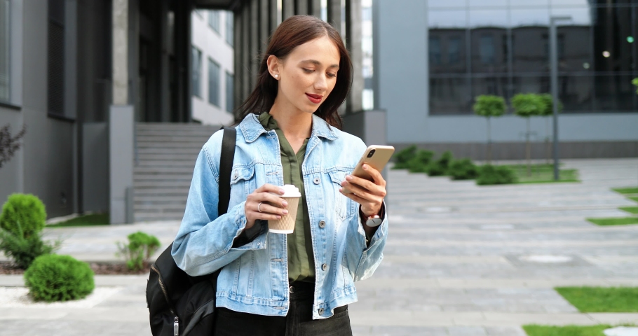 Caucasian joyful young stylish woman tapping or scrolling on smartphone and sipping hot drink in the morning at street. Beautiful happy female texting message on phone and drinking coffee. Outside. | Shutterstock HD Video #1059904682