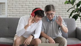 Asian father and son waving hands while wearing headphones looking at camera, make video call with digital laptop with family in the living room. Their are chatting and relaxing with family.