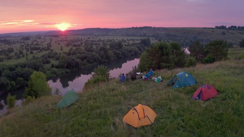 Camping family aerial. Extended family sit on the coast of the river with hiking camps set around and enjoy gentle sunset.
