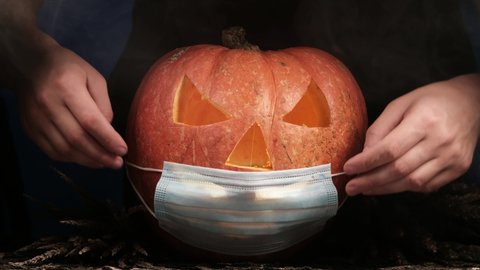 male hands dress up for Halloween carved pumpkin glowing with protective medical mask on face, coronavirus and quarantine concept, covid-19 halloween costume.