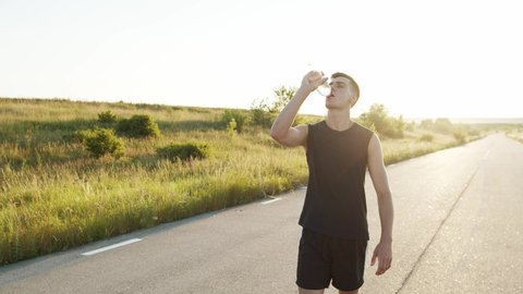 Young sportsman walking on the road at summer nature with sunset and drinking water from a bottle. Slowly in 4K