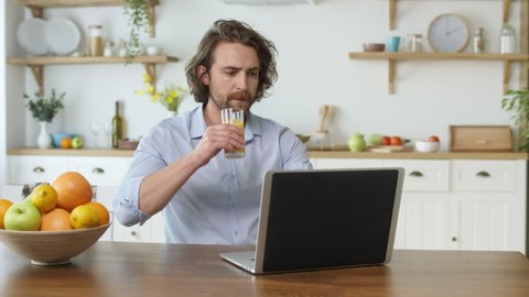 Attractive Man Working With A Laptop From Home. Man With Mustache and Long Hair Sitting at the Table Drink a Juice From the Glass During Remotely Work From Home.