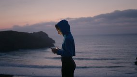 Side View of Young Beautiful Woman Standing Oceanfront and Typing on Smartphone. Female Traveler Posting Photos of Astonishing Ocean Landscape on Social Media. Travel Concept. Slow Motion Shot