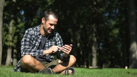 Young handsome guy in a plaid shirt leafs through the feed of social networks while sitting on the grass in the park. A young guy reads the feed of social networks on his smartphone while sitting on