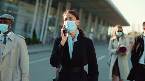 Young businesswoman walking with crowd of office people talking smartphone wearing protective face masks going to work outdoors. Social distancing. Quarantine.
