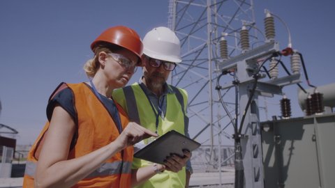 Low angle of mature man and woman in vests and helmets browsing tablet while working on modern power plant