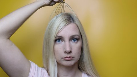 A caucasian blond woman enjoy head scalp massage by anti stress acupuncture metal octopus tool, equipment, she closes her eyes with pleasure, yellow background.