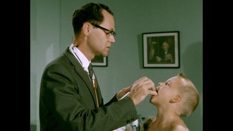 1950s: Little boy sits on doctors examination table. Medium shot of Doctor puts a tongue depressor in the boys mouth and examines his throat. Close-up of boy being examined. Doctor uses stethoscope.
