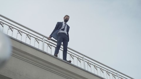 Extreme wide shot of frustrated Caucasian businessman in Covid-19 face mask attempting to commit suicide. Bottom view portrait of male bankrupt standing on bridge ready to jump down.