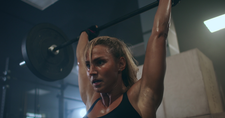 A female weightlifter performs a barbell lift in a dark gym. a woman lifting a heavy bar over her head | Shutterstock HD Video #1059957179