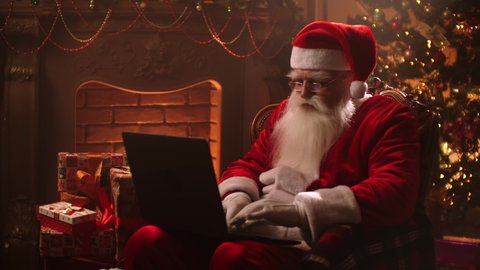 Modern Santa Claus. Cheerful Santa Claus working on laptop and smiling while sitting at his chair with fireplace and Christmas Tree in the background.