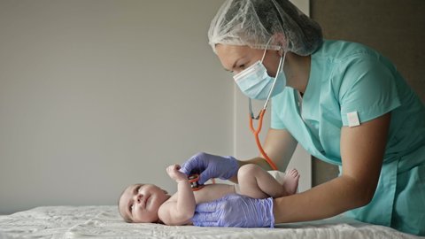 Neonatologist wearing latex gloves and a medical mask listens to a newborn baby with a stethoscope.