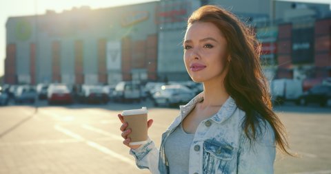 A woman is standing with a cup of coffee in the parking lot of a shopping center. She is smiling. 4K